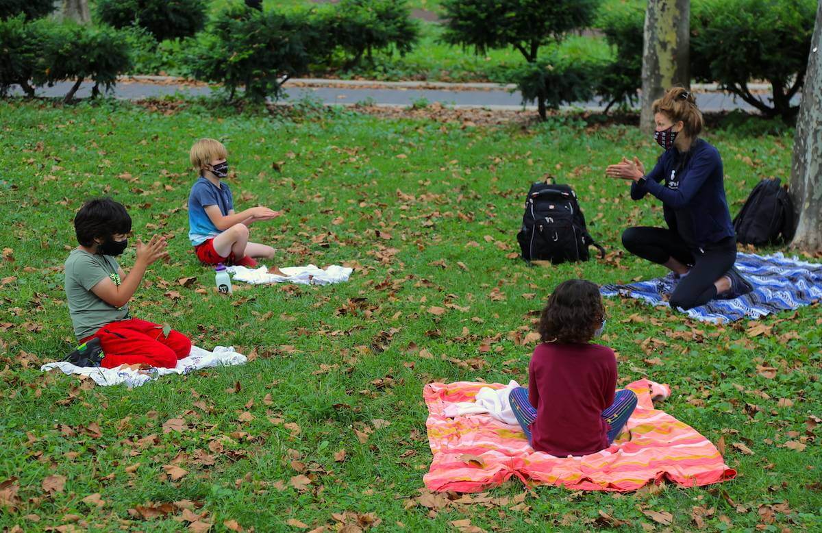 Outdoor class with teacher and kids sitting outside on blankets on grass, all wearing face masks